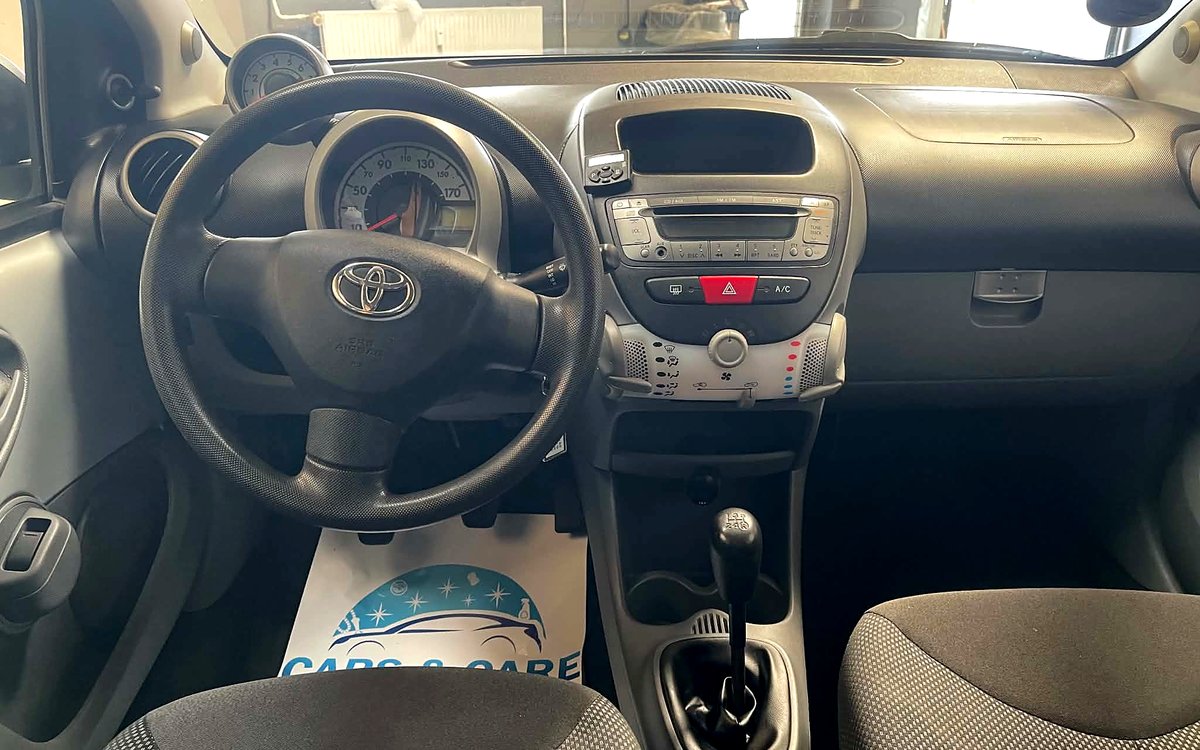 Instrumentbrættet i den sølvgrå Toyota Aygo med rat, radio, aircondition og manuelt gear