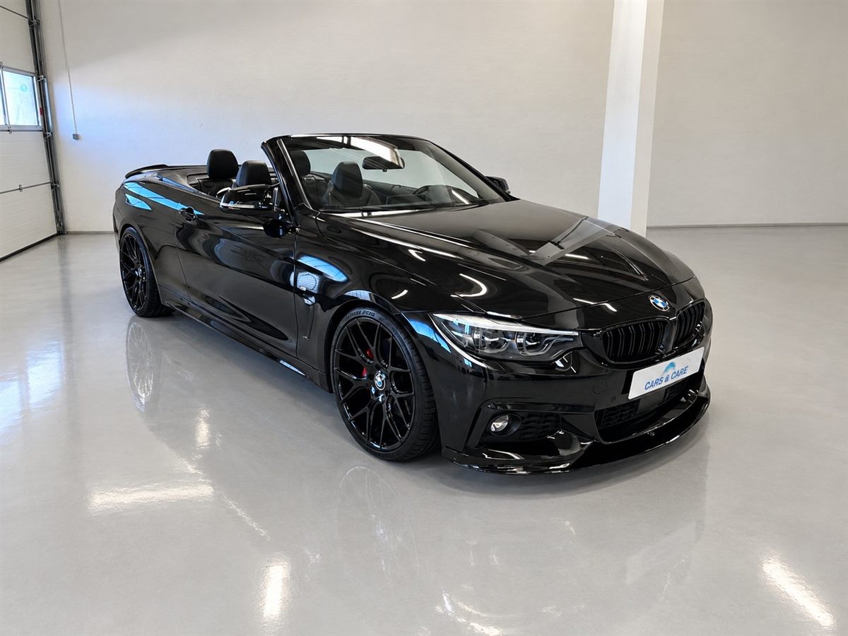 Blank, sort BMW-cabriolet med sorte fælge og Cars & Care-nummerplade på det lyse værkstedsgulv, hvor bilen spejler sig