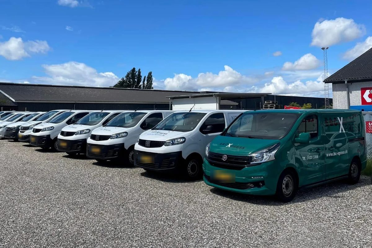 En lang række hvide varevogne og en grøn varevogn parkeret på en grusplads