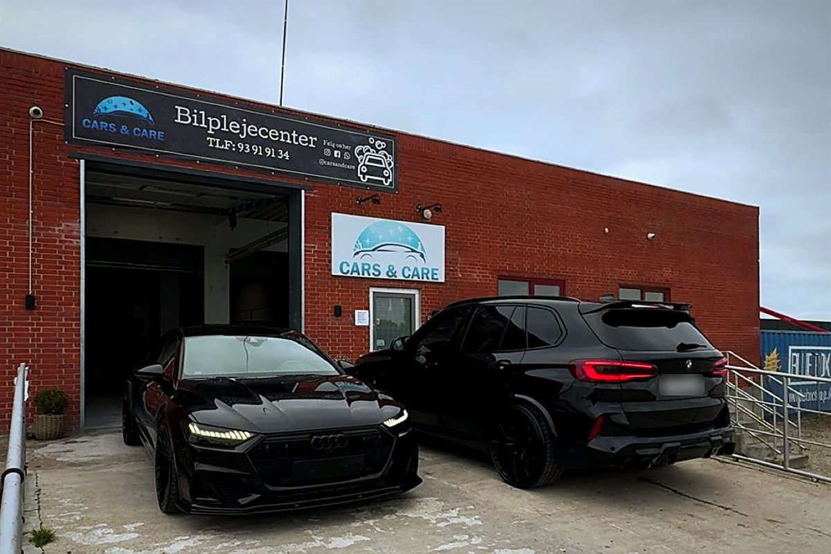 Værkstedets røde murstensfacade med skiltet Cars & Care Bilplejecenter, den åbne port og to sorte biler på pladsen foran
