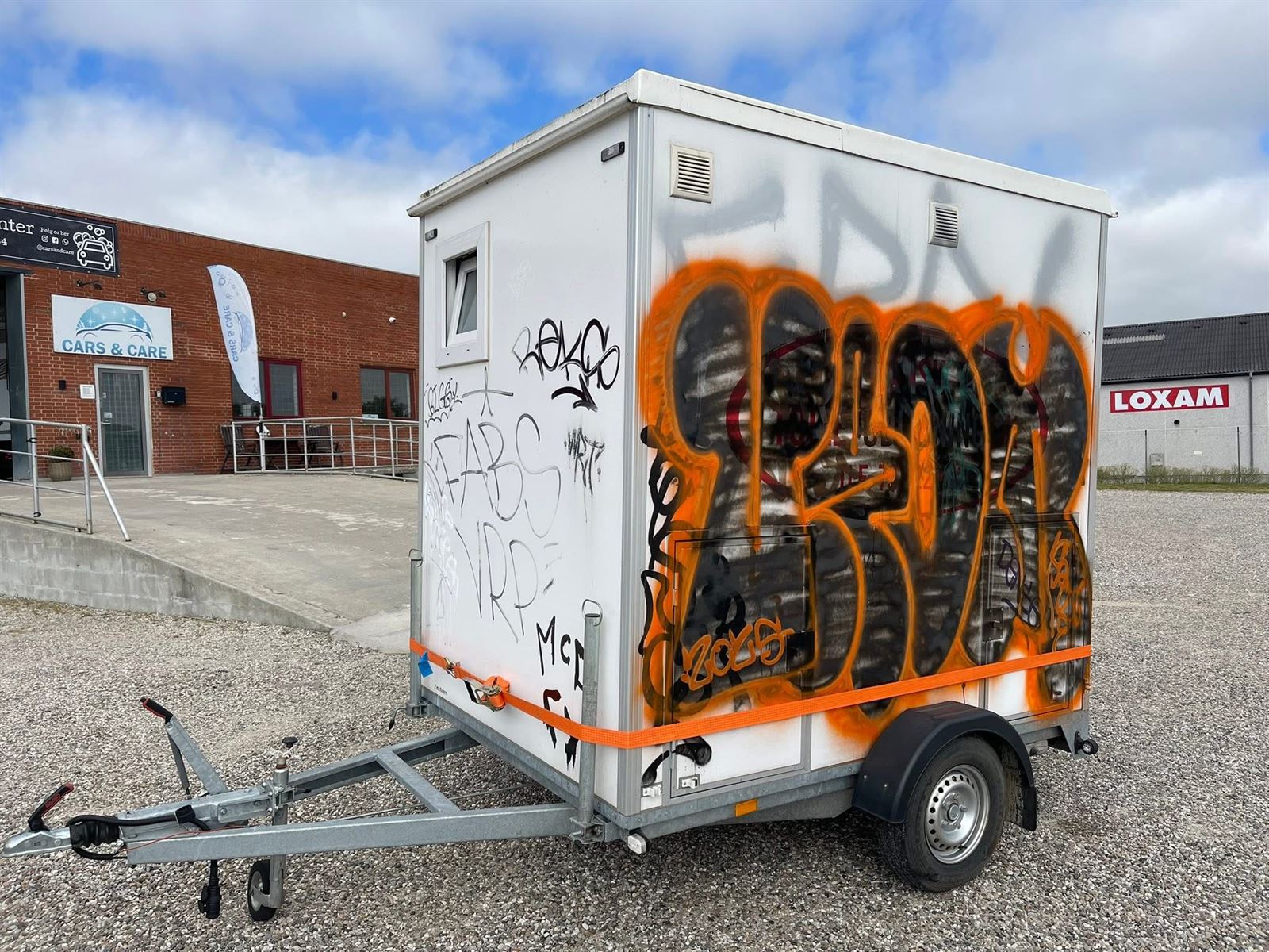Hvid lukket trailer dækket af orange og sort graffiti foran værkstedet